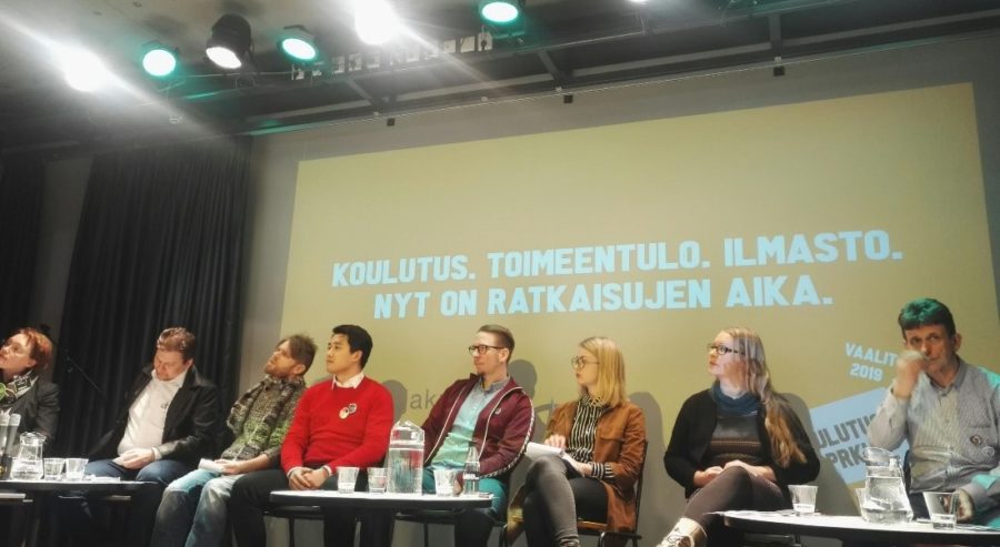 Vaalipaneelin osallistujat istuvat rivissä lavalla. Taustalle on heijastettu paneelinaihe, "Koulutus. Toimeentulo. Ilmasti. Nyt on ratkaisujen aika."