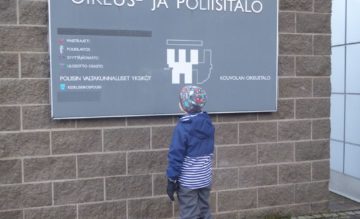 Lapsi seisoo selin kameraan oikeus- ja poliisitalon opastekyltin edessä.