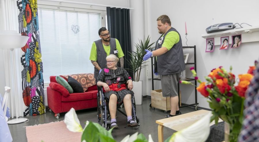 Kaksi kotihoidon työntekijää seisoo olohuoneessa keskustelemassa. Asiakas istuu pyörätuolissa ja seuraa keskustelua.